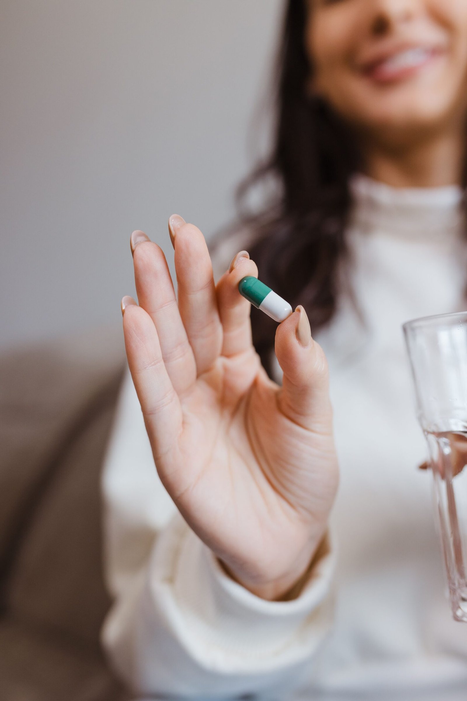 Smiling woman holds a green and white capsule with a glass of water indoors.